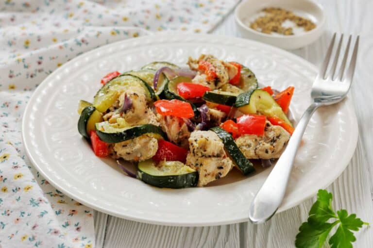 A white plate with Italian herbed chicken and veggie bake foil packs, a fork on the side, and a small dish of spices in the background.