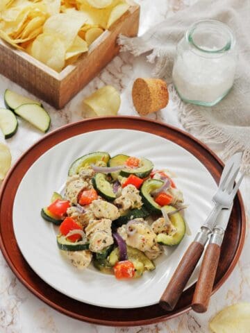 A plate of Italian Herbed Chicken and Veggie Bake Foil Packs is served with a side of potato chips and a jar of salt on a marble table.