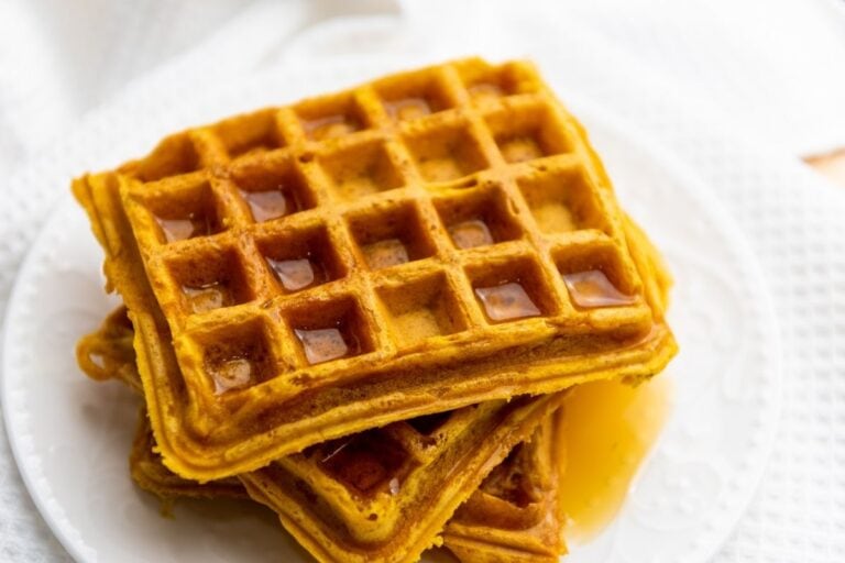 A stack of square waffles with syrup on a white plate, placed on a white textured surface.