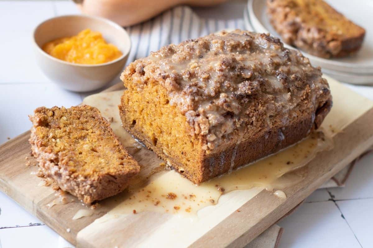 A loaf of pumpkin bread with streusel topping and glaze sits on a wooden board, with one slice cut and a bowl of pumpkin puree nearby.