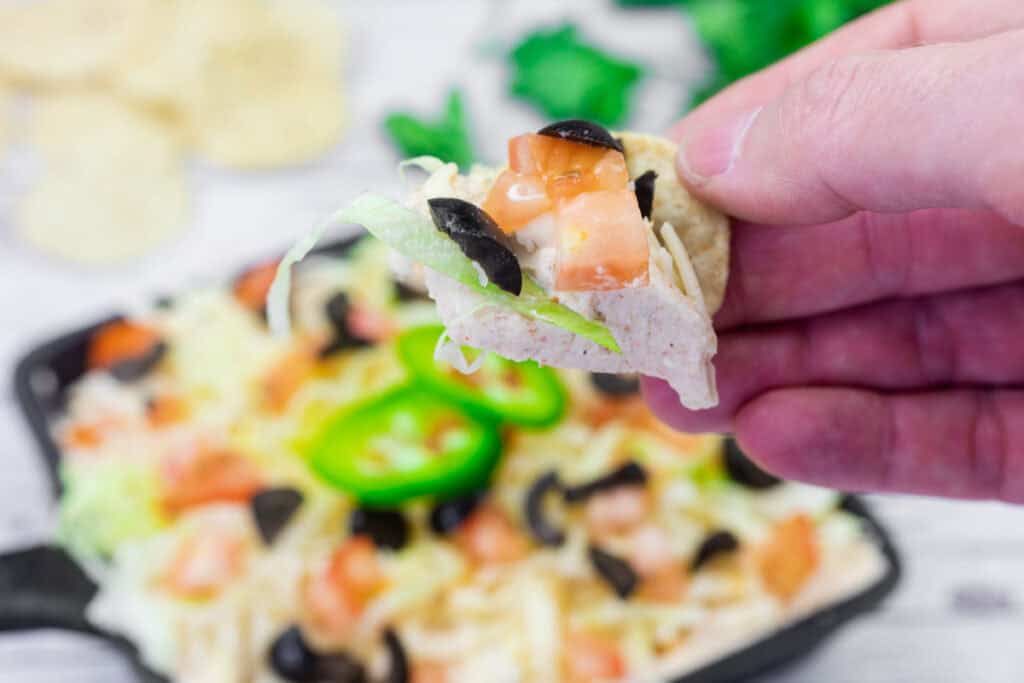 A hand holds a tortilla chip topped with lettuce, tomato, cheese, and black olives above a plate of nachos.