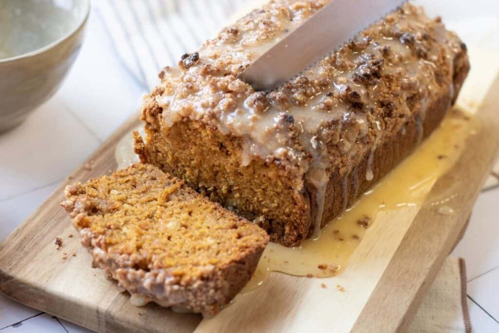 A loaf of iced pumpkin bread is being sliced on a wooden cutting board, with one slice cut and glaze dripping onto the board.