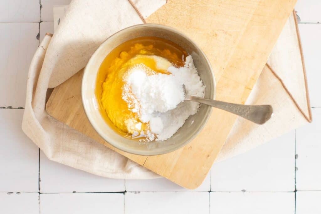 A bowl with egg yolks and sugar being mixed with a spoon, placed on a wooden board and a beige cloth on a tiled surface.