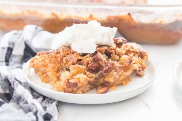 A slice of Pumpkin Crunch Cake served on a white plate with a dollop of whipped cream, napkin beside it, and a baking dish in the background.