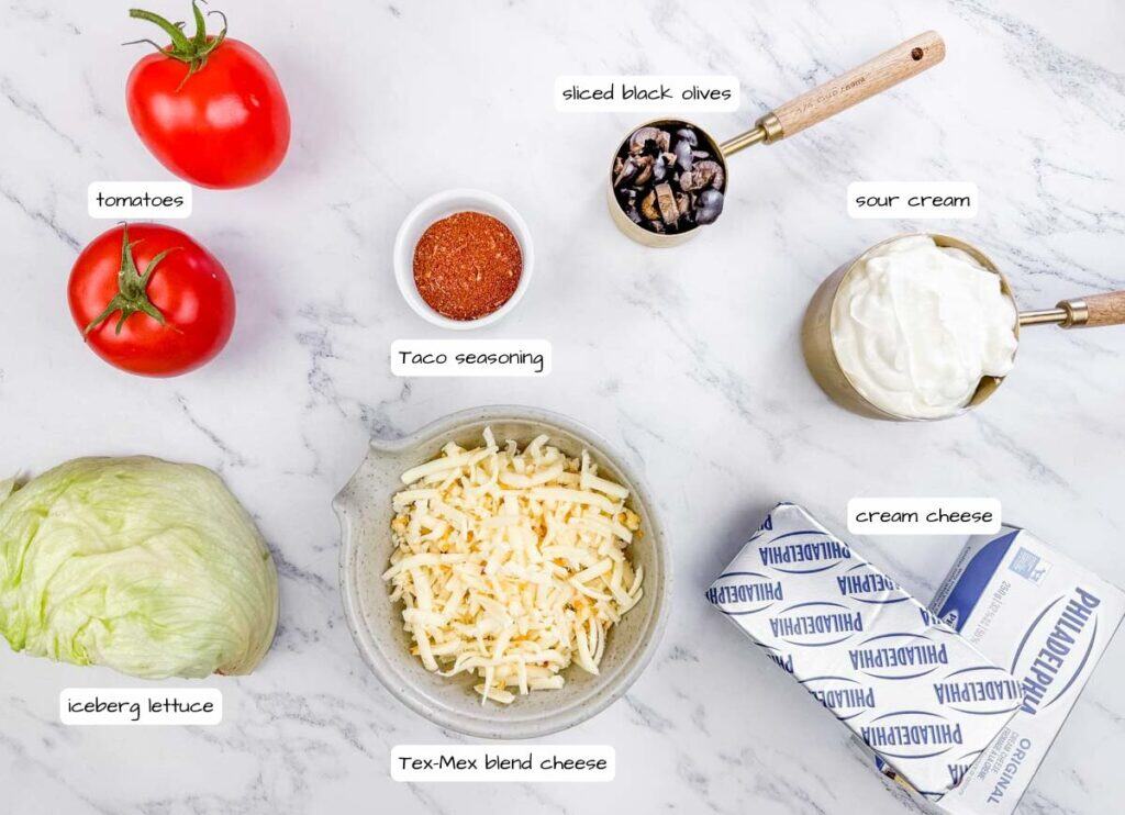 Ingredients for a dish laid out on a marble surface: tomatoes, taco seasoning, sliced black olives, sour cream, cream cheese, Tex-Mex blend cheese, and iceberg lettuce.