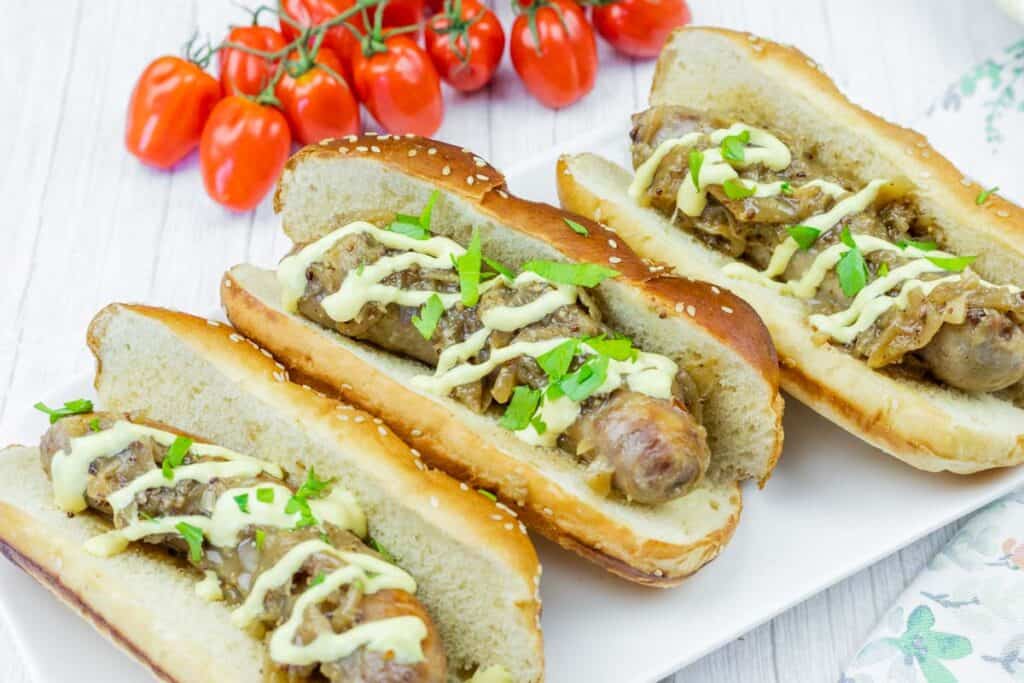 Three sausage sandwiches in buns topped with grilled onions, drizzled with sauce, and garnished with parsley. A bunch of cherry tomatoes is in the background.