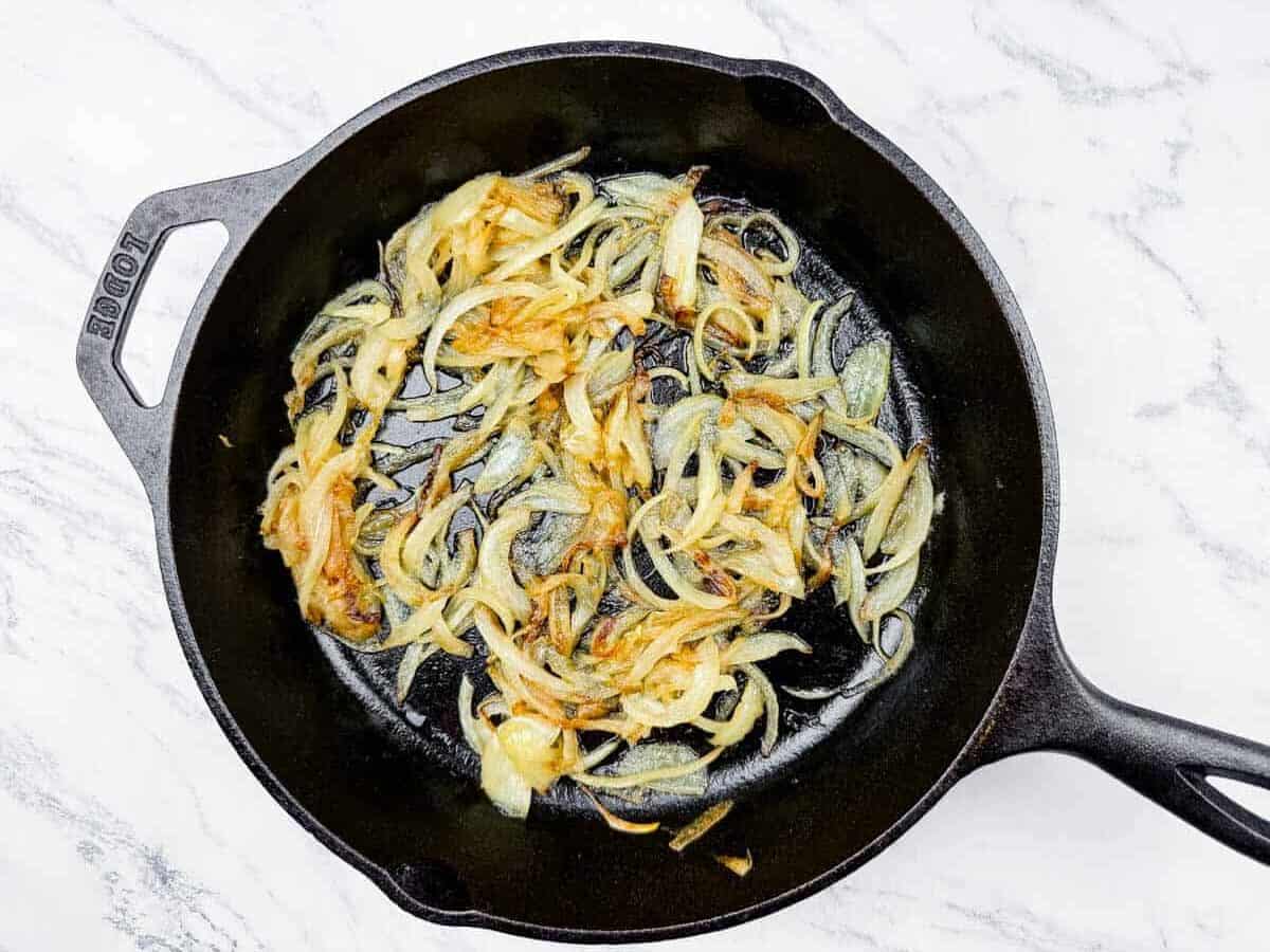 Sliced onions being saut&eacute;ed and caramelized in a black cast iron skillet on a white marble surface.