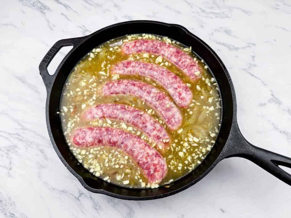 A cast iron skillet with five raw sausages, chopped garlic, and liquid on a marble countertop.