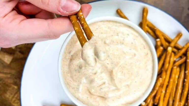 A hand dips two pretzel sticks into a bowl of creamy dip, with additional pretzel sticks on the plate beside the bowl.