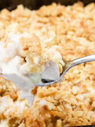 A hand holds a spoonful of ritz cracker chicken casserole with cheese, with the casserole dish in the background.