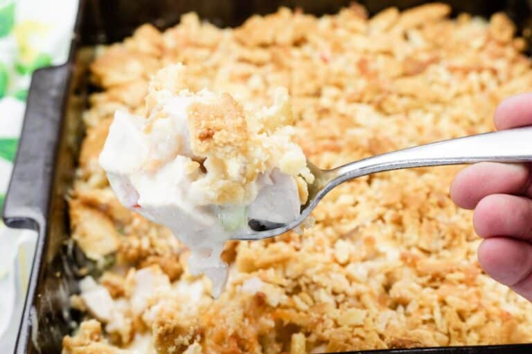 A hand holds a spoonful of ritz cracker chicken casserole with cheese, with the casserole dish in the background.