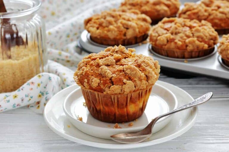 A muffin with crumb topping sits on a white plate with a spoon, next to a jar of brown sugar and a muffin tray on a light wooden surface.