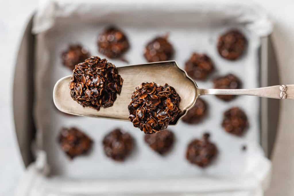 A metal spatula holds two chocolate oat cookies above a baking tray lined with parchment paper and filled with more cookies.