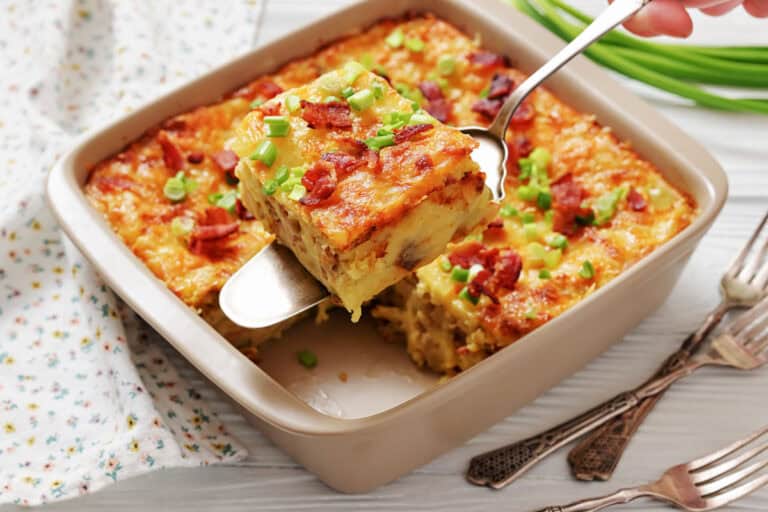 A baked casserole with cheese, bacon, and green onions is being served with a spatula from a square baking dish.