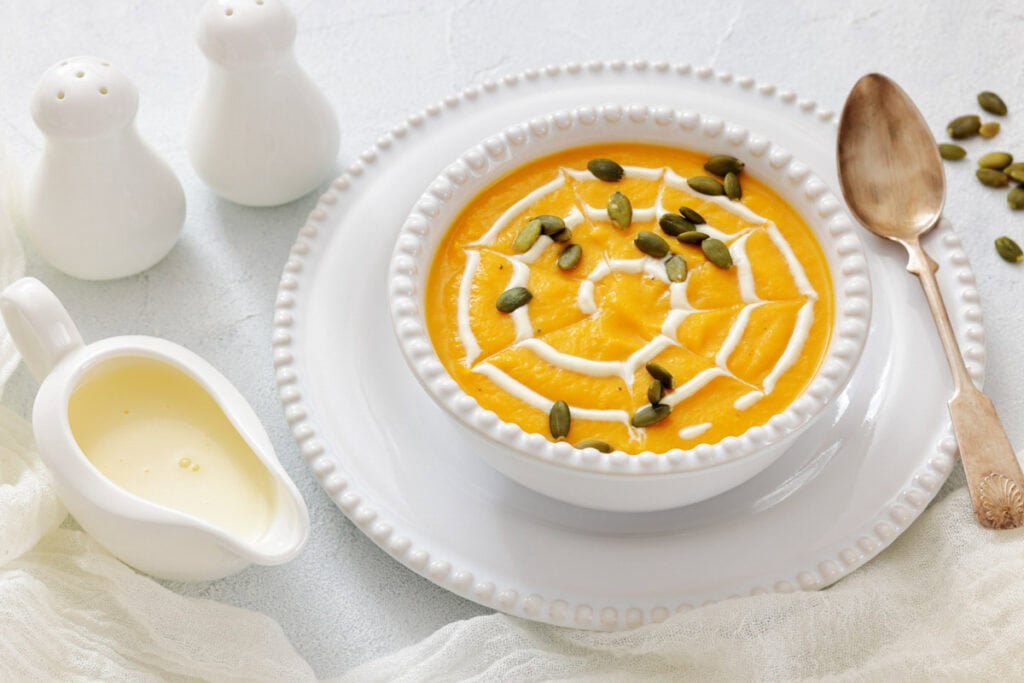 A bowl of pumpkin soup garnished with cream and pumpkin seeds, served on a white plate with a spoon and cream jug on the side. Salt and pepper shakers are in the background.