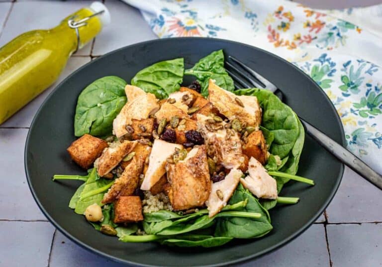 A black plate with spinach, grilled chicken pieces, roasted sweet potatoes, nuts, and seeds, with a fork on the side and a bottle of yellow dressing in the background.