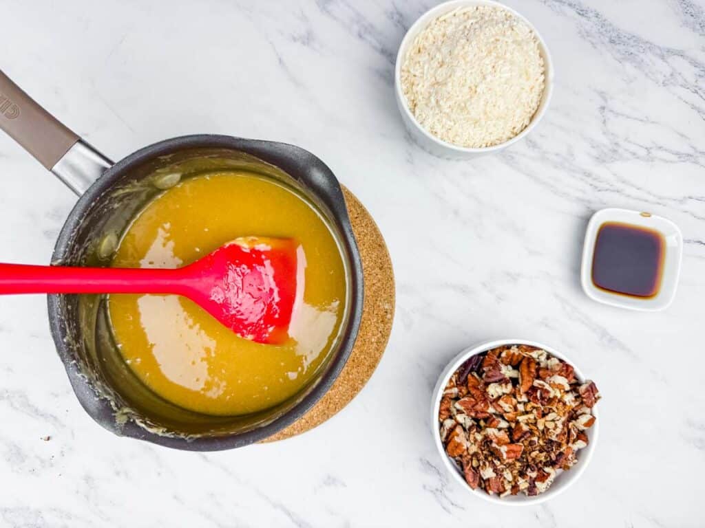 A saucepan with a red spatula stirring a caramel-like mixture, surrounded by bowls of shredded coconut, chopped pecans, and a small dish of vanilla extract on a marble surface.