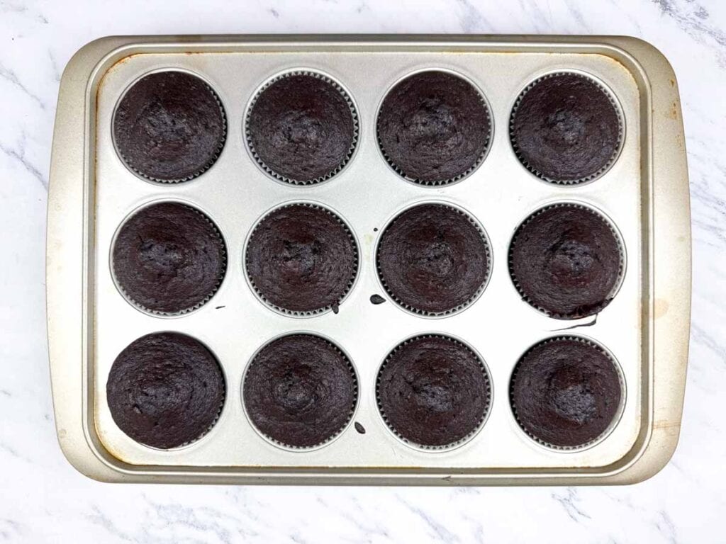 A metal muffin tray holds twelve baked chocolate cupcakes in paper liners, viewed from above on a white marble surface.