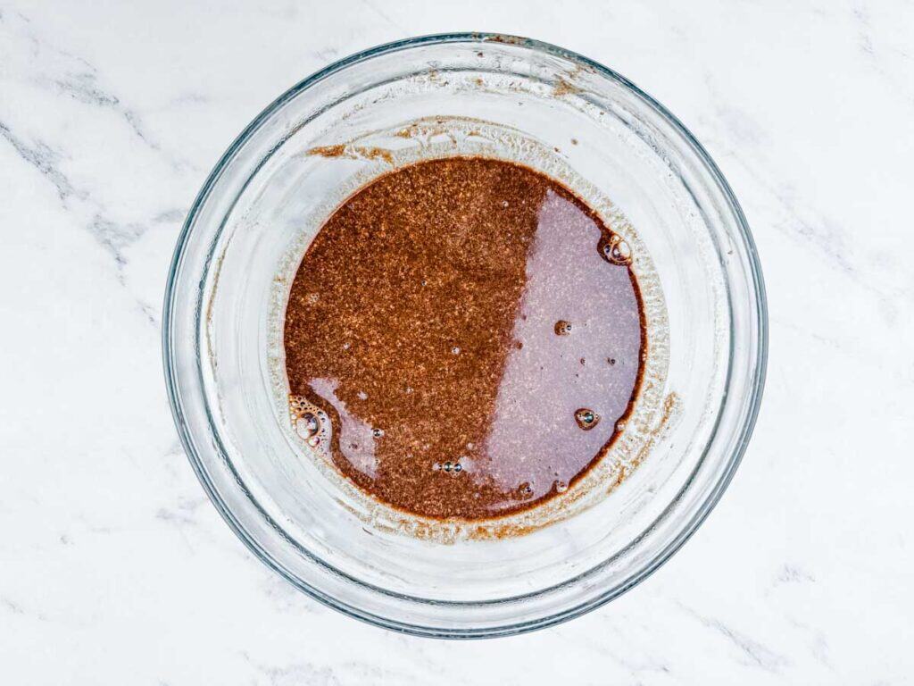 A clear glass bowl containing a mixture of buttermilk and melted chocolate chips, placed on a white marble surface.