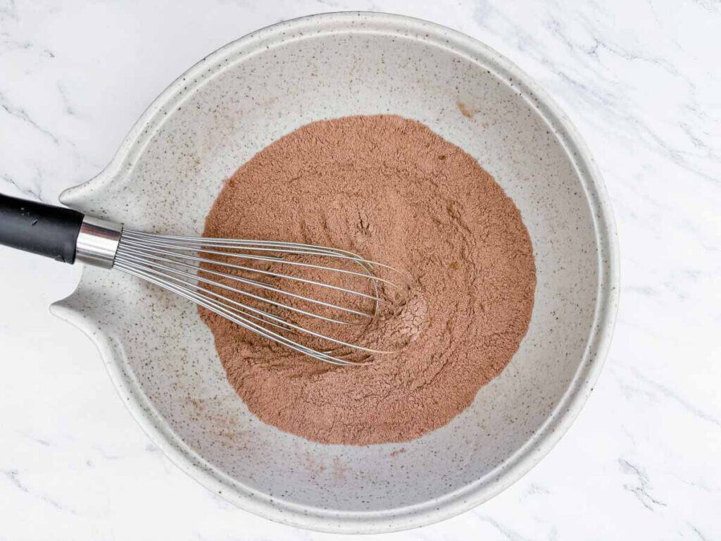 A metal whisk mixing dry ingredients in a large speckled ceramic bowl on a white marble surface.