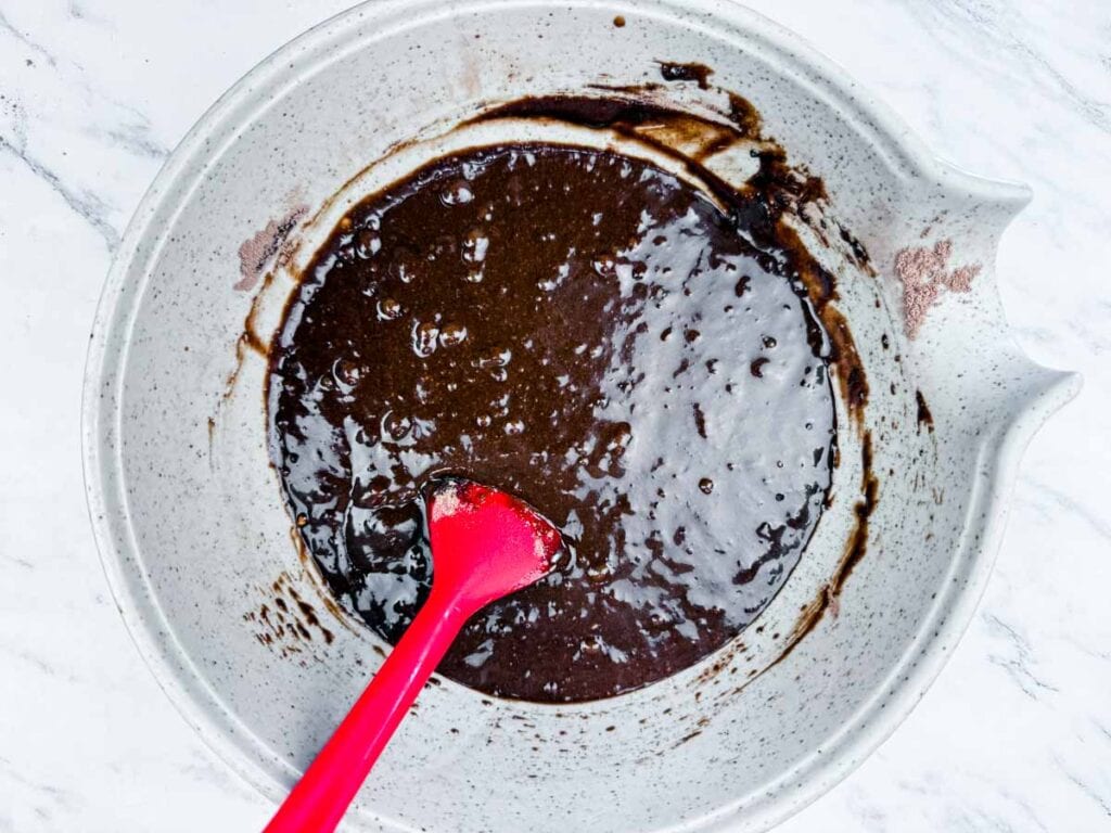 A mixing bowl containing chocolate batter being stirred with a red spatula on a white marble surface.