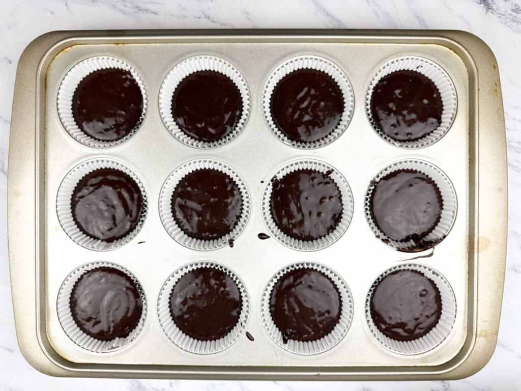 A muffin tin filled with twelve paper cupcake liners, each containing chocolate batter, on a marble countertop.