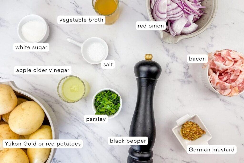Ingredients for a dish are arranged on a white countertop, including potatoes, bacon, red onion, vegetable broth, sugar, salt, mustard, vinegar, parsley, and black pepper.