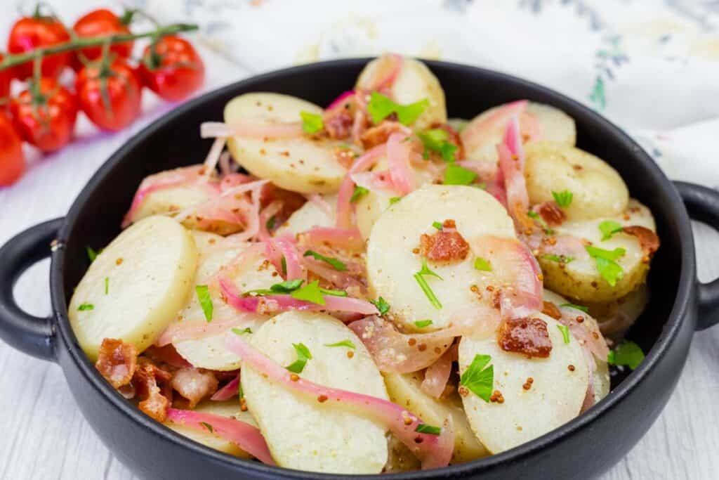 A black bowl filled with sliced potatoes, red onions, bacon pieces, chopped parsley, and whole grain mustard. Cherry tomatoes are in the background.