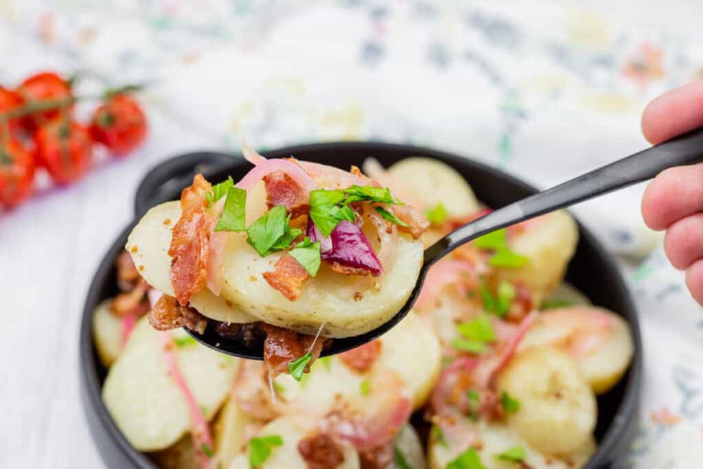 A spoonful of German potato salad topped with bacon, red onion, and chopped parsley held over a bowl of the same salad.