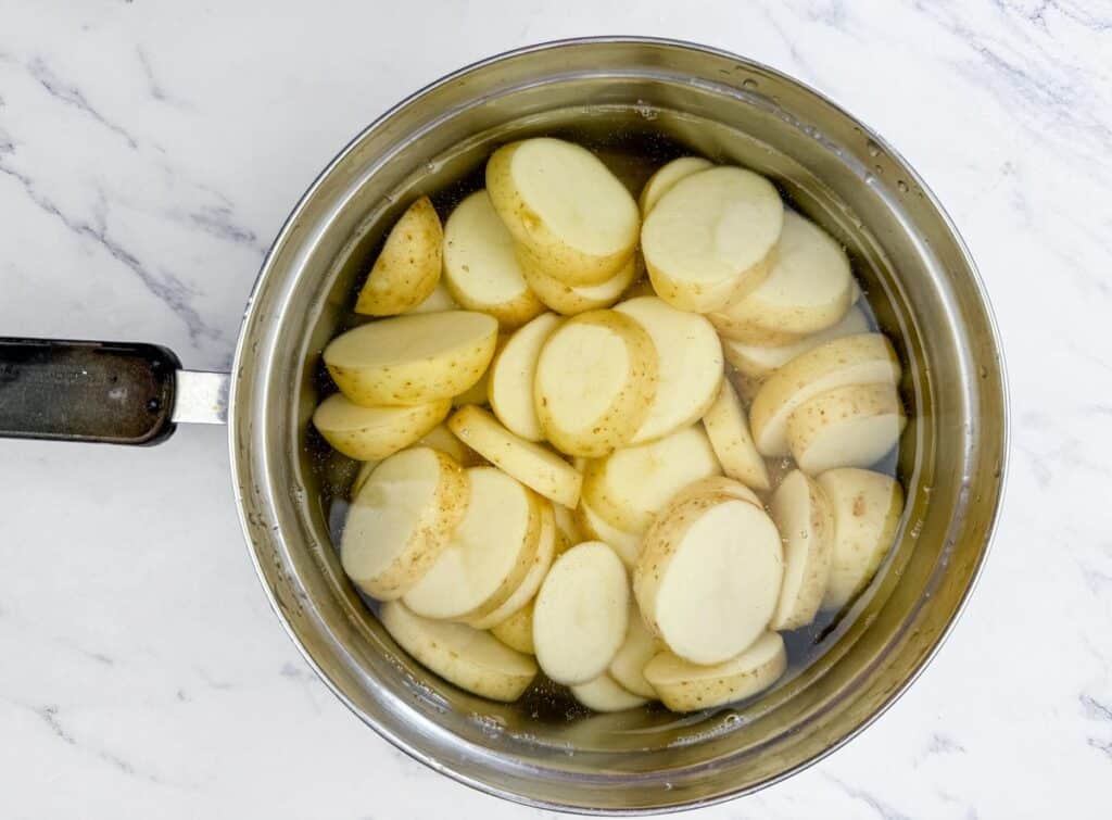 Sliced potatoes in water inside a stainless steel pot, placed on a white marble surface.