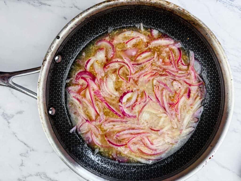 A frying pan on a marble surface containing sliced red onions cooking in oil and liquid.