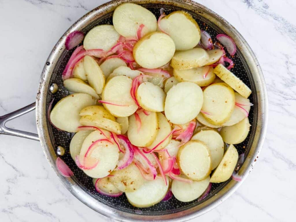 Sliced potatoes and red onions cooking in a stainless steel skillet on a white marble countertop.
