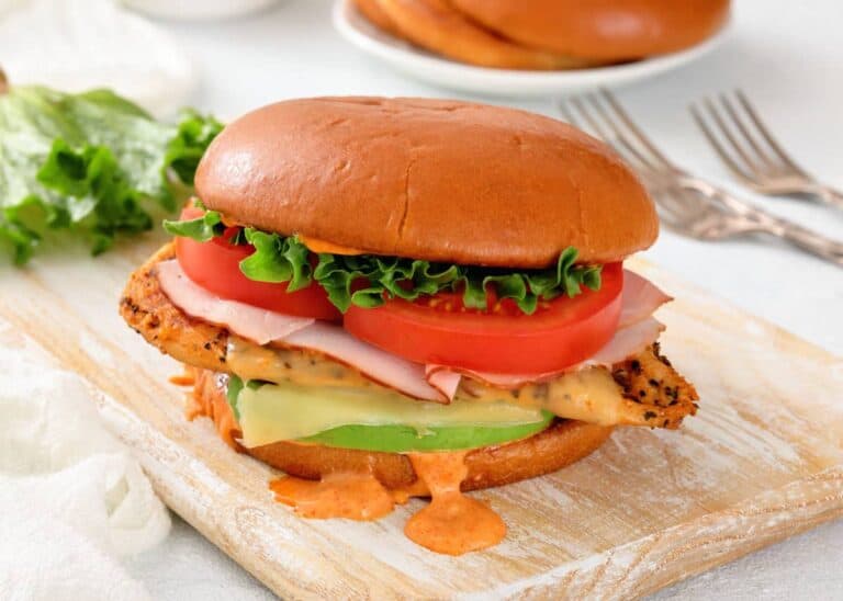 A sandwich with a grilled chicken breast, ham, cheddar cheese, lettuce, tomato, avocado, and sauce on a toasted bun sits on a wooden board.