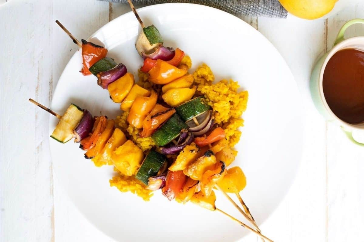 Three Grilled Teriyaki Mango Skewers with bell peppers, zucchini, and onions served on a bed of yellow rice on a white plate.