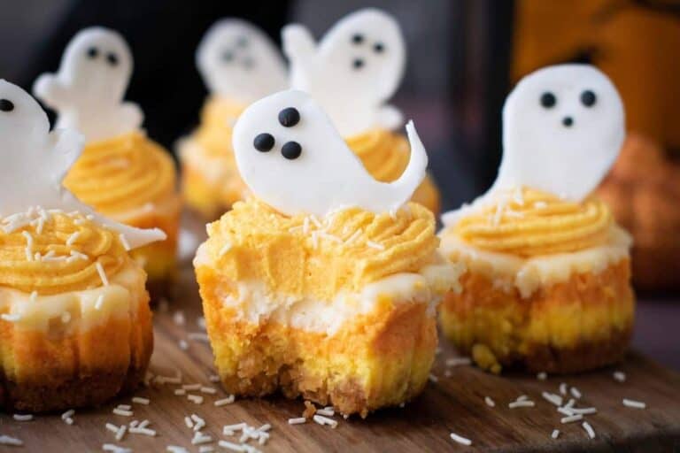 Mini cheesecakes with yellow and orange frosting, topped with white ghost-shaped decorations and chocolate eyes, sit on a wooden surface with sprinkled white shreds.
