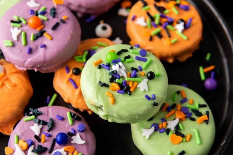 Three colorful cookies coated in purple, orange, and green icing are decorated with Halloween-themed sprinkles and arranged on a dark plate.