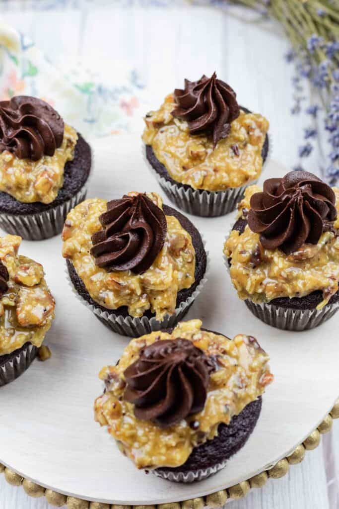 Six chocolate cupcakes topped with caramel, coconut, and pecan frosting, each finished with a swirl of chocolate frosting, displayed on a round white tray.