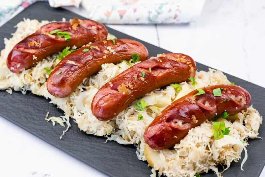 Four grilled sausages served on a bed of sauerkraut and sliced potatoes, garnished with chopped parsley, on a black rectangular plate.