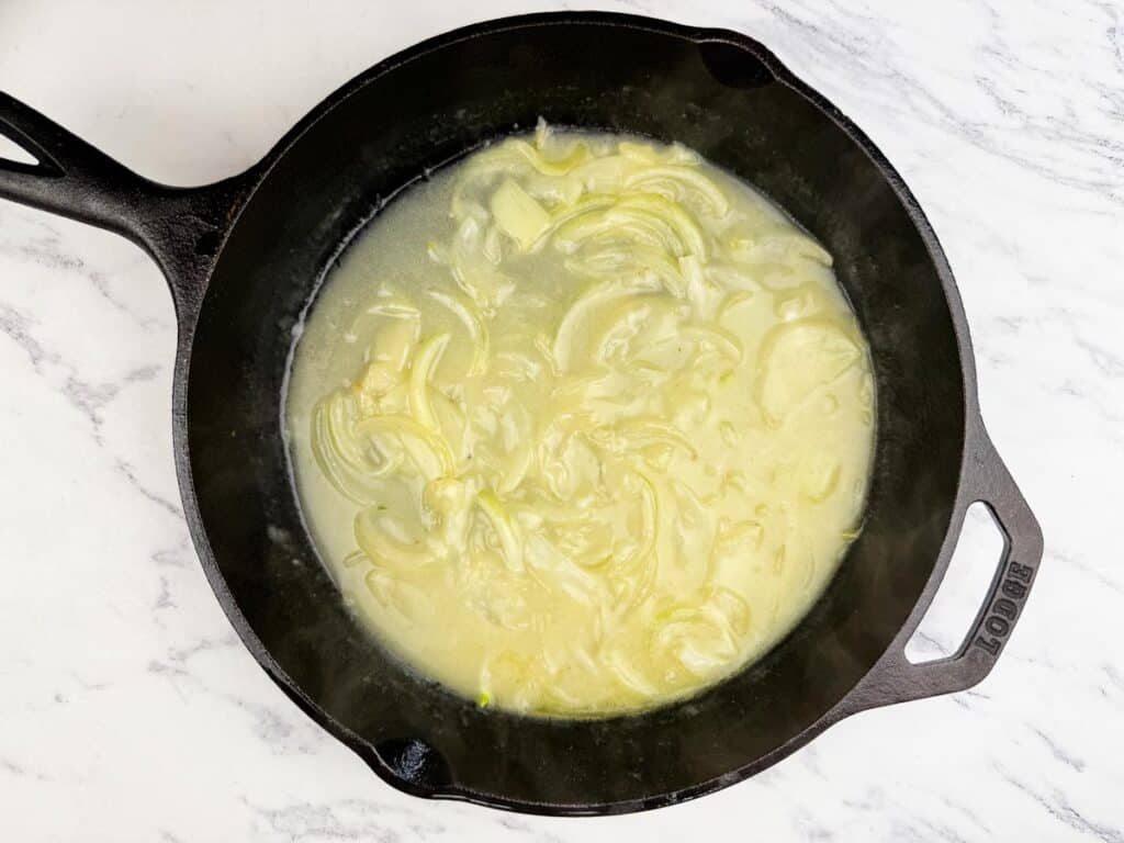 Sliced onions cooking in a cast iron skillet with liquid, set on a white marble surface.