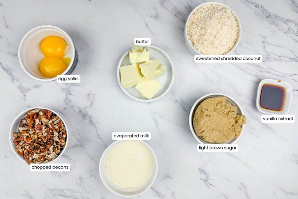Six labeled ingredients&mdash;egg yolks, butter, sweetened shredded coconut, chopped pecans, evaporated milk, light brown sugar, and vanilla extract&mdash;arranged on a marble surface.