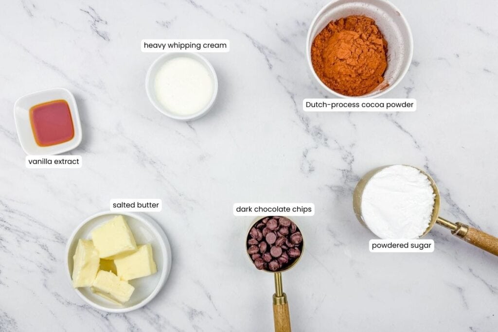 Six labeled baking ingredients&mdash;heavy whipping cream, Dutch-process cocoa powder, vanilla extract, salted butter, dark chocolate chips, and powdered sugar&mdash;arranged on a white marble surface.
