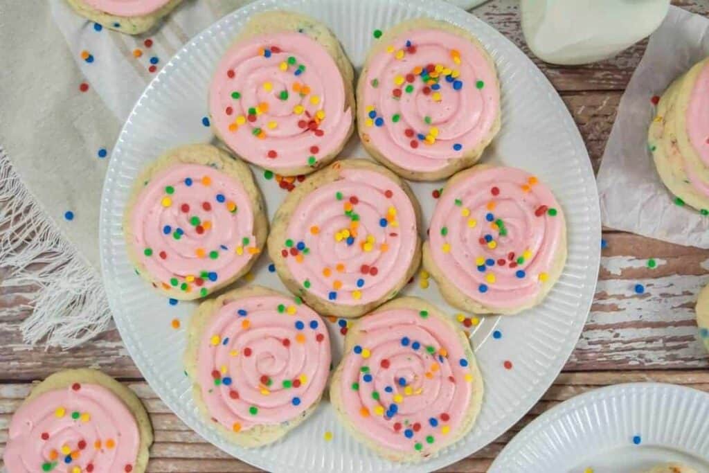 A plate of Copycat Crumbl Confetti Cake Cookies topped with pink frosting and multicolored sprinkles sits on a wooden table.