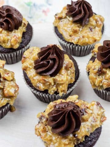 Six chocolate cupcakes topped with a layer of pecan-coconut caramel and a swirl of chocolate frosting, arranged on a white round tray.