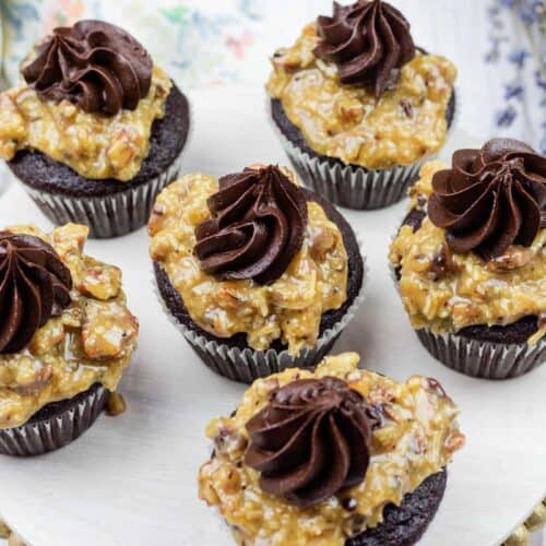 Six chocolate cupcakes topped with a layer of pecan-coconut caramel and a swirl of chocolate frosting, arranged on a white round tray.