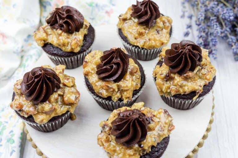 Six chocolate cupcakes topped with a layer of pecan-coconut caramel and a swirl of chocolate frosting, arranged on a white round tray.