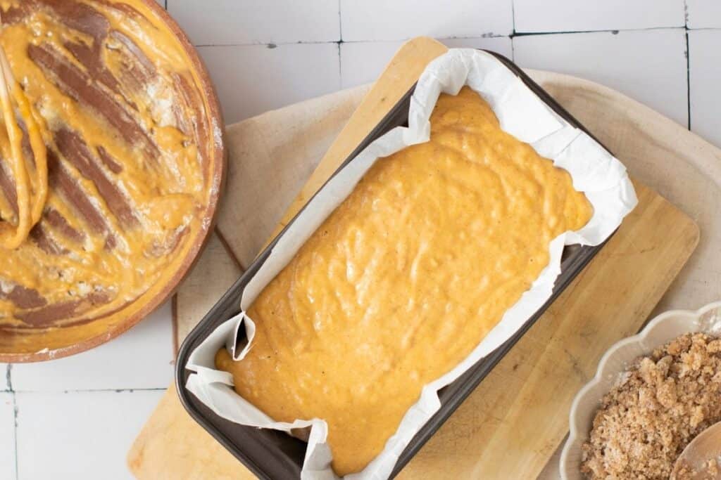 A loaf pan lined with parchment paper filled with unbaked batter sits on a cutting board beside a mixing bowl with leftover batter and a small bowl of crumb topping.