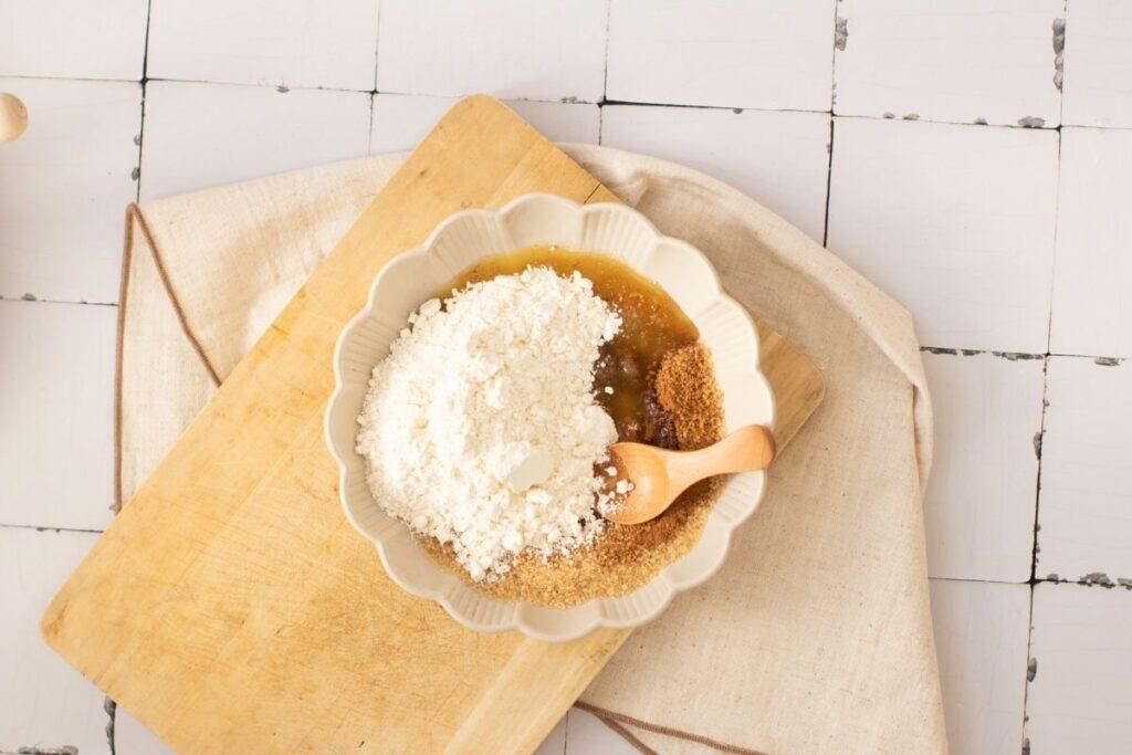 A white bowl on a wooden cutting board contains flour, brown sugar, and a liquid mixture, with a small wooden spoon resting inside.