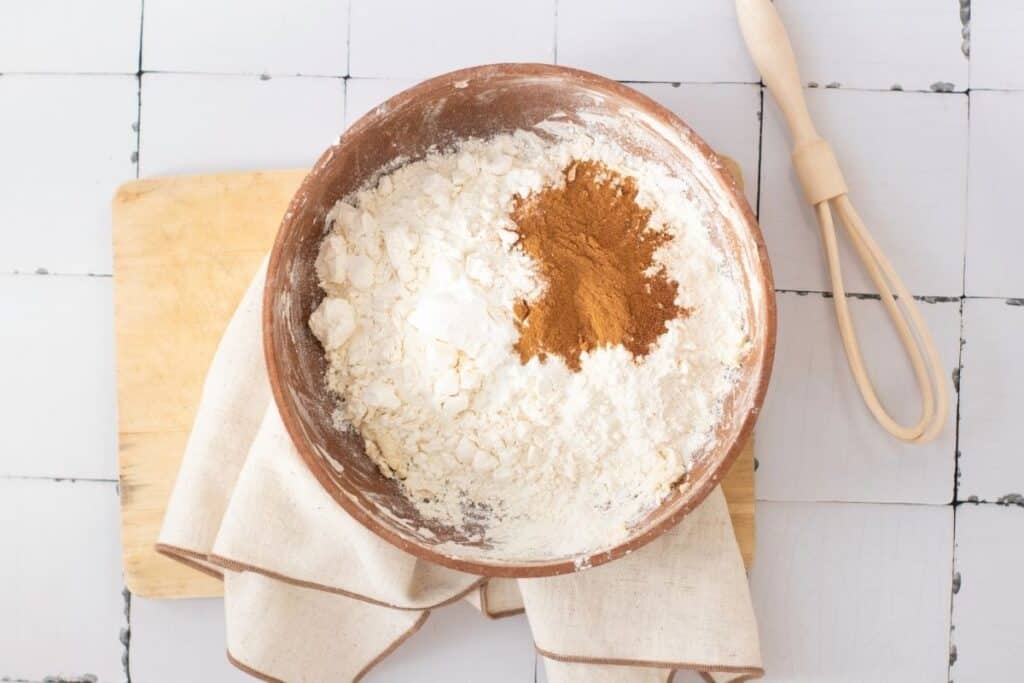 A bowl with flour and ground cinnamon sits on a cloth, next to a whisk and wooden board on a tiled surface.