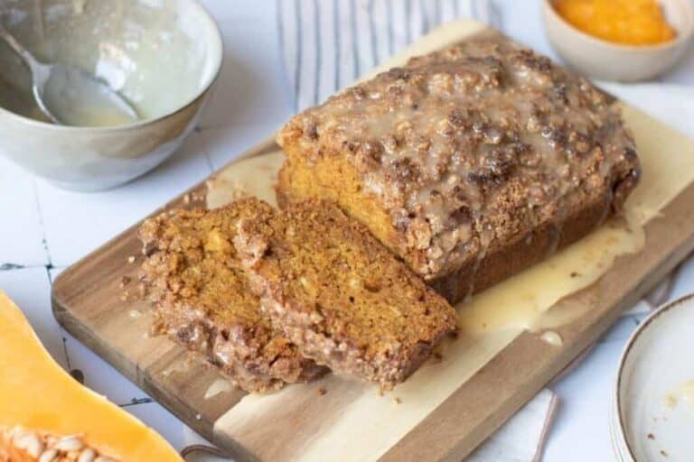 A loaf of glazed pumpkin bread with a crumb topping sits on a wooden board, with two slices cut and placed in front.