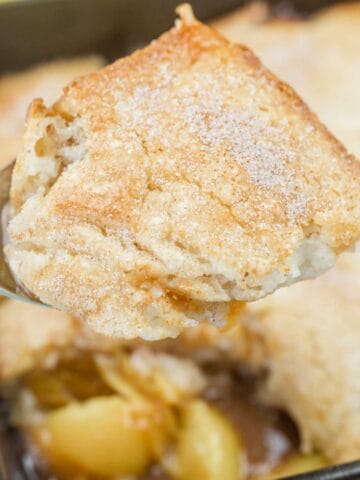 A close-up of a serving of apple cobbler with a golden, sugar-dusted biscuit topping and visible apple filling underneath.